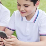 happy schoolchildren using smartphone together while sitting on grass
